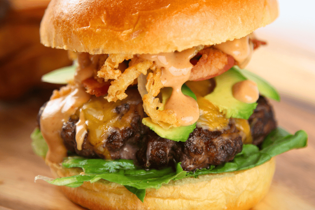 A cheeseburger with lettuce, avocado slices, crispy onions, bacon, and sauce in a toasted bun.