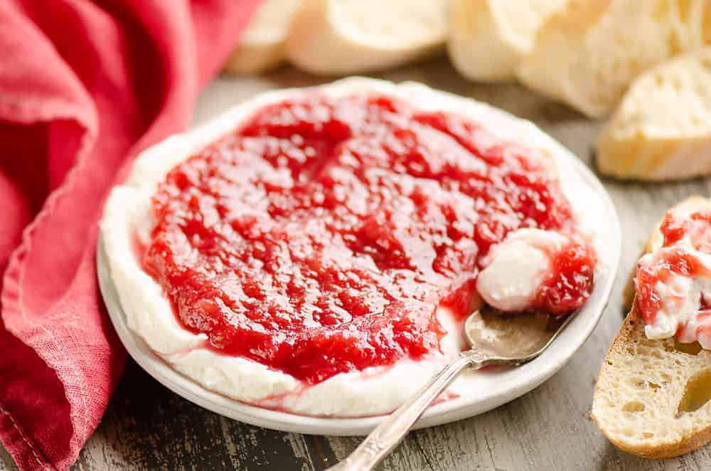 A plate of whipped cream cheese topped with strawberry jam, with a spoon resting on the plate and slices of bread in the background.