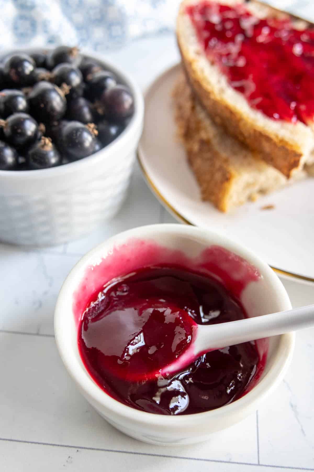 A small bowl of glossy blackcurrant jam with a spoon, a bowl of blackcurrants, and a plate with bread spread with jam in the background.