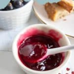 A white bowl filled with black currant jelly and a spoon, with black currants and bread slices in the background.