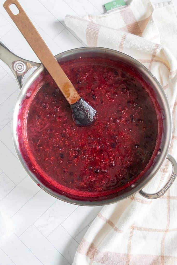 A saucepan of mixed berry compote with a black spatula on a white tiled surface next to a beige and white checkered towel.