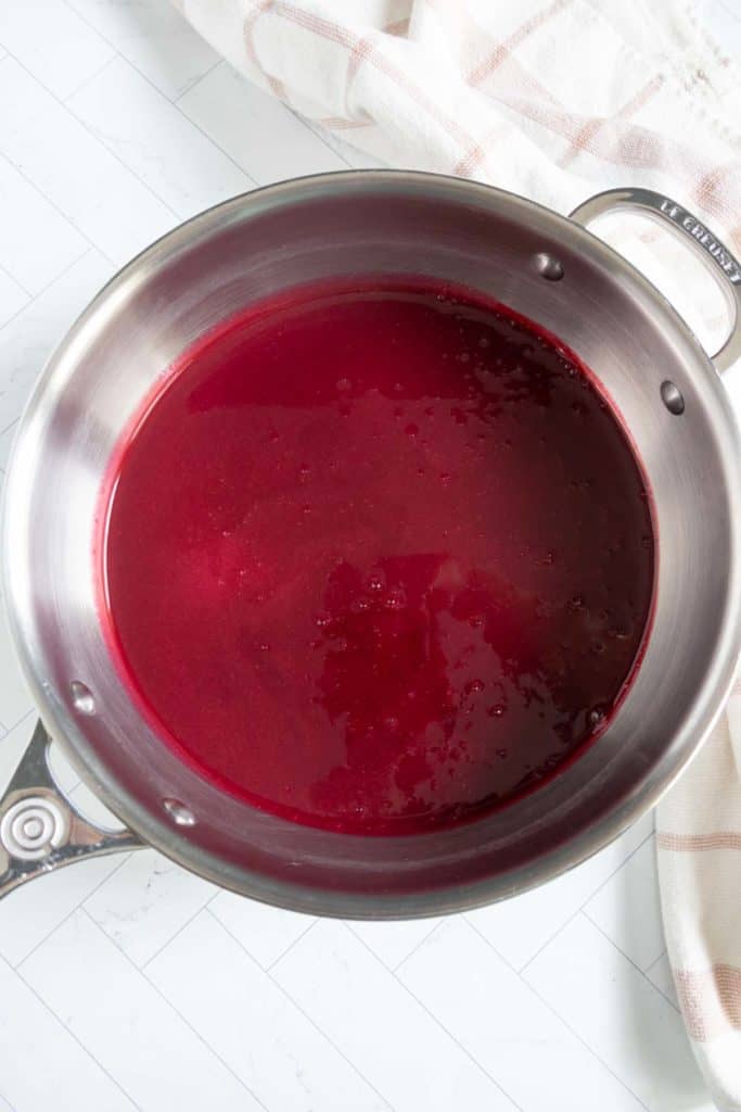 A stainless steel pot filled with a deep red liquid sits on a white countertop next to a beige and red striped cloth.