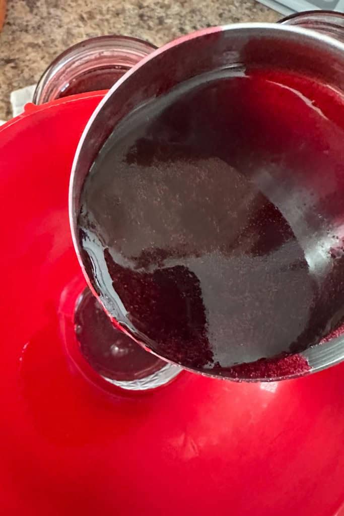 A pot of dark purple liquid is being poured into a glass jar through a red funnel on a kitchen countertop.