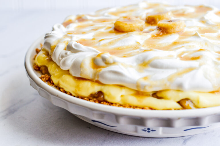 A banana cream pie in a white pie dish, topped with whipped cream, caramel drizzle, and banana slices.