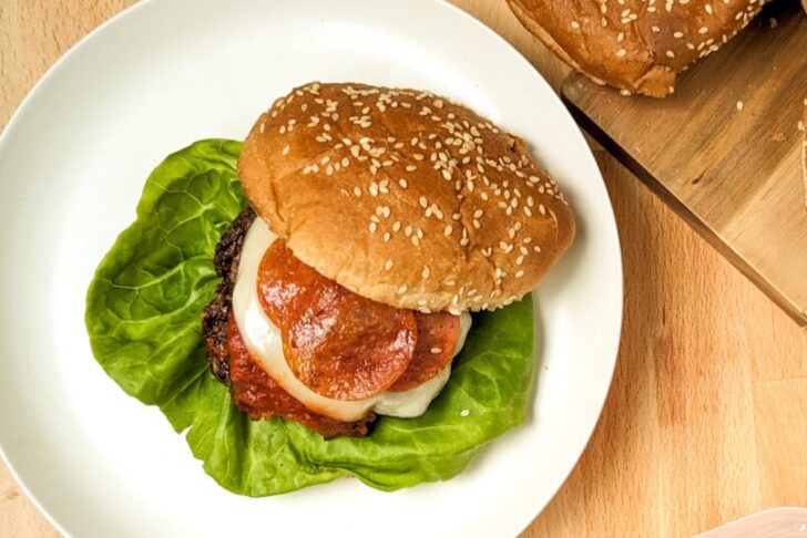 A hamburger with lettuce, cheese, pepperoni, and sauce on a sesame seed bun, served on a white plate.