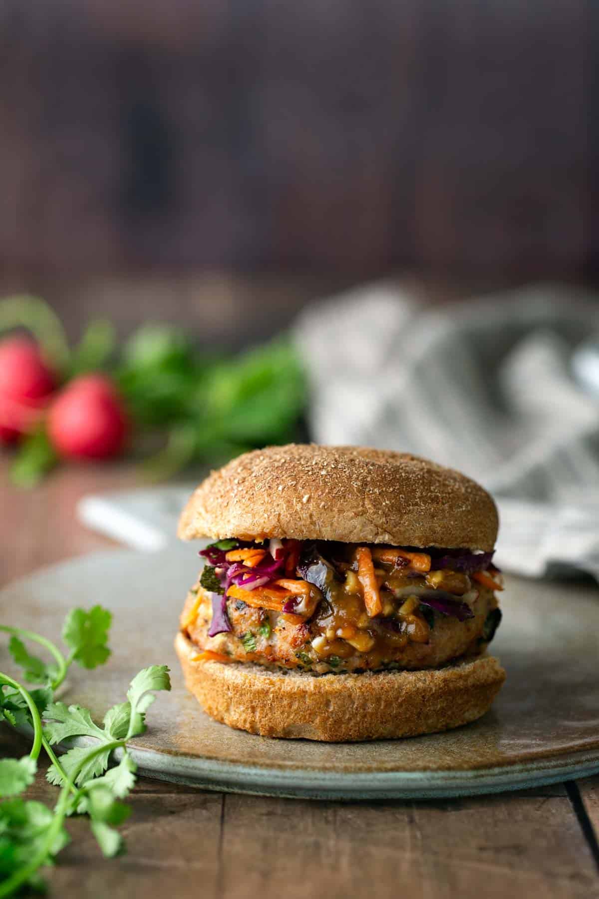 A sandwich with a vegetable patty and colorful slaw on a whole wheat bun sits on a ceramic plate, with herbs and radishes in the background.