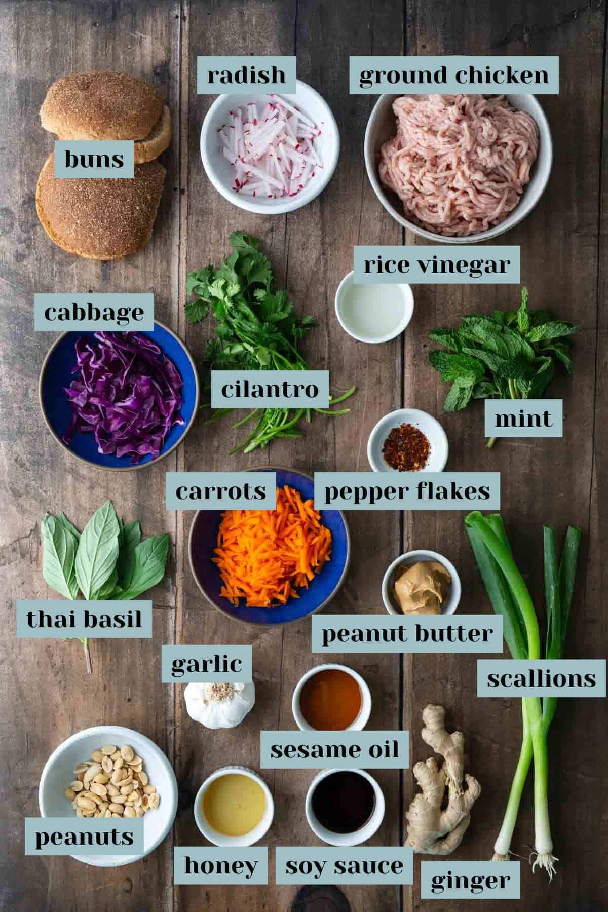 Various labeled ingredients for a ground chicken dish, including vegetables, herbs, sauces, buns, peanuts, and seasonings, arranged on a wooden surface.