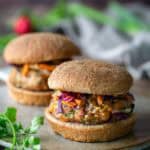 Two veggie burgers with whole wheat buns, topped with shredded vegetables and sauce, sit on a ceramic plate with herbs nearby.