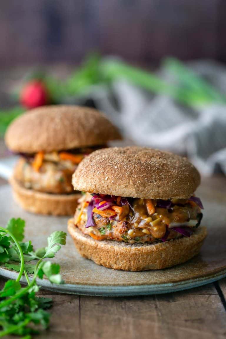 Two veggie burgers with whole wheat buns, topped with shredded vegetables and sauce, sit on a ceramic plate with herbs nearby.
