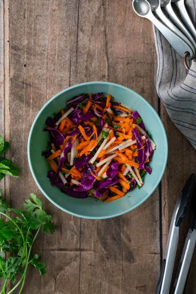A bowl of shredded purple cabbage, carrots, and white vegetables on a wooden table, with herbs, tongs, and measuring spoons nearby.