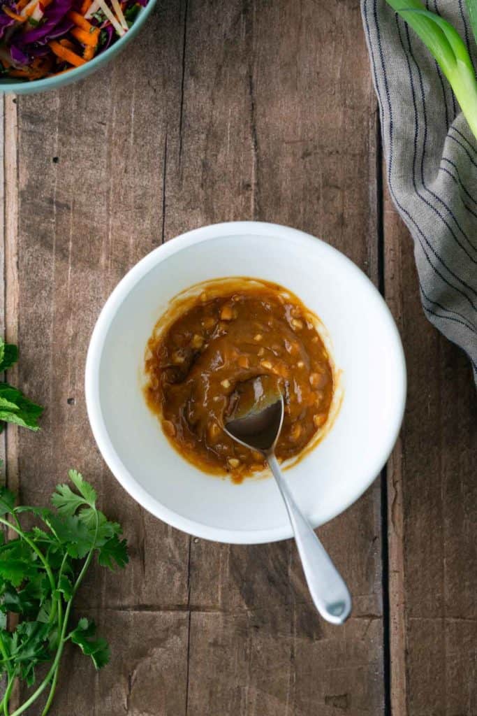 A white bowl containing a brown sauce with a spoon, placed on a wooden surface, surrounded by fresh herbs and a striped towel.
