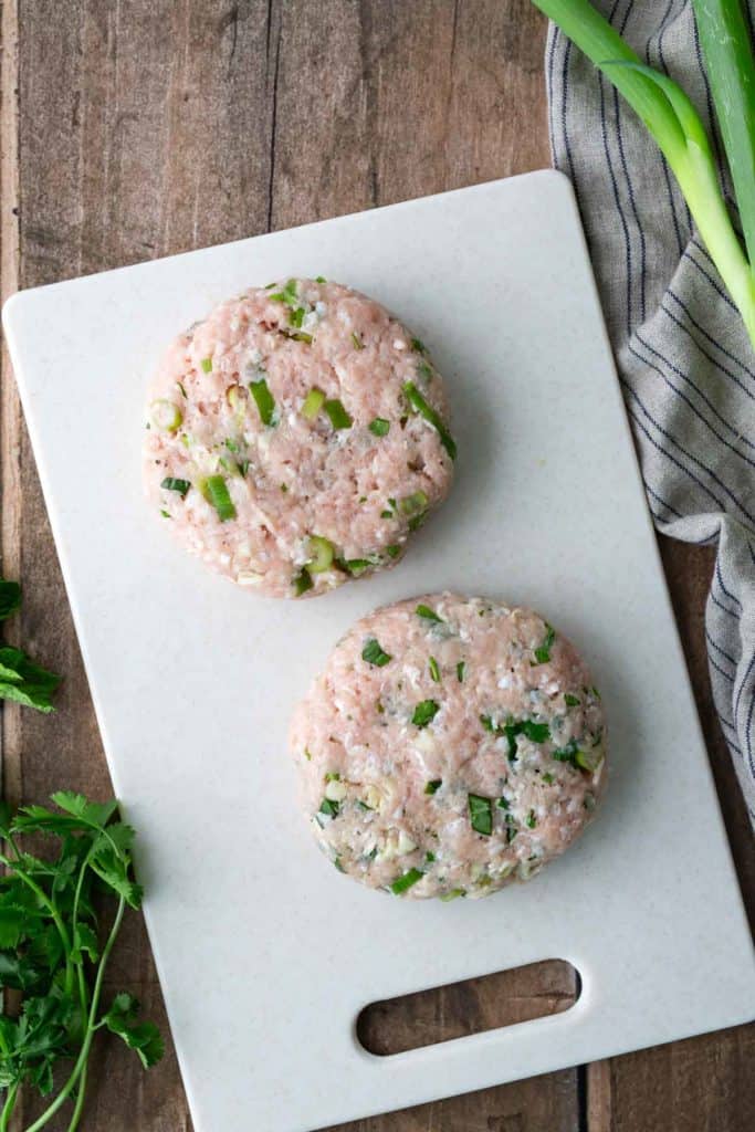 Two raw chicken burger patties with chopped green onions and herbs on a white cutting board, next to fresh cilantro and a striped kitchen towel.