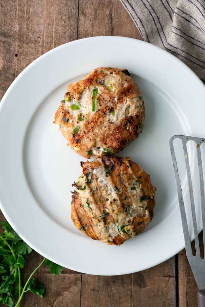 Two grilled turkey burger patties with herbs on a white plate, next to a metal spatula and a sprig of parsley on a wooden surface.