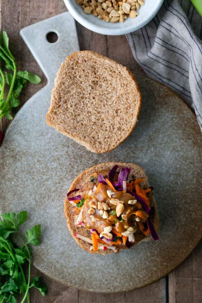 An open-faced sandwich with shredded vegetables and chopped peanuts on a whole wheat bun, placed on a round gray cutting board.