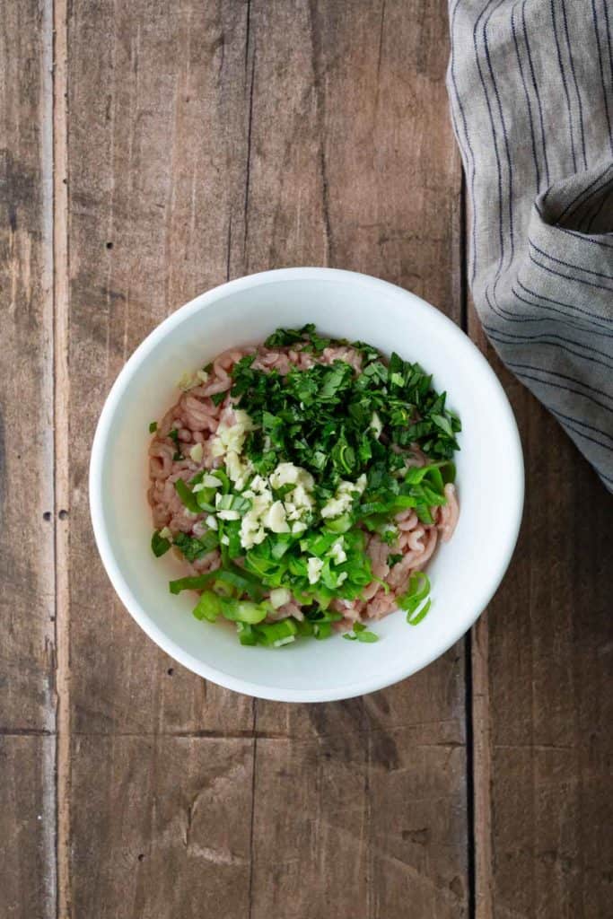 A white bowl containing ground meat, chopped green onions, minced garlic, and fresh herbs sits on a wooden surface next to a striped cloth.