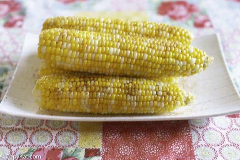 Four ears of cooked corn on the cob with seasoning are stacked on a white plate, placed on a colorful patterned tablecloth.