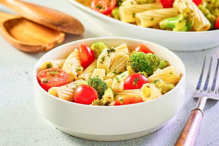 A white bowl filled with rigatoni pasta, cherry tomatoes, and broccoli, garnished with herbs. A fork and wooden utensils are nearby.