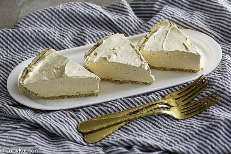 Three slices of creamy pie with graham cracker crust on a white plate, set on a striped cloth beside three gold forks.