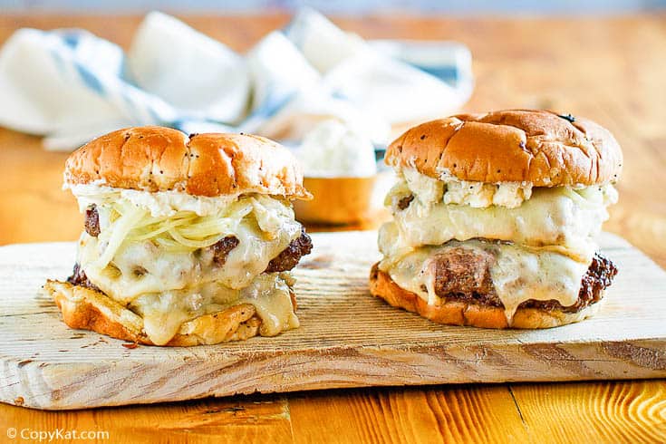 Two cheeseburgers with melted cheese, grilled onions, and a creamy topping sit on a wooden board.