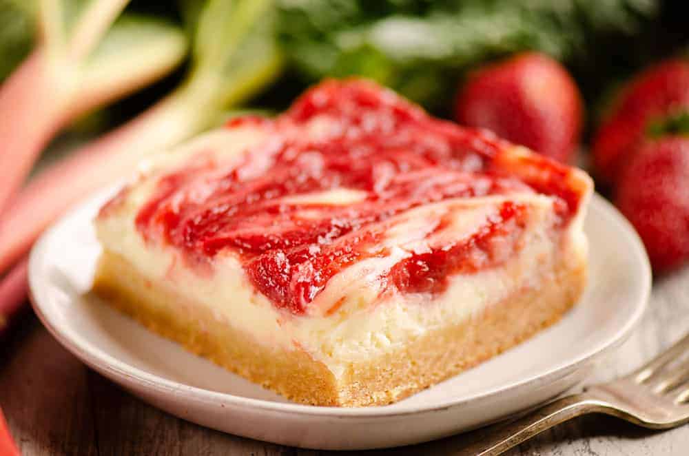 A square slice of strawberry cheesecake bar with a swirled strawberry topping on a white plate, with strawberries and greenery in the background.
