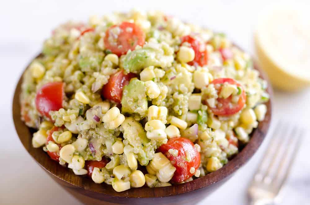 A wooden bowl filled with a salad of corn, cherry tomatoes, avocado, red onion, and quinoa, with a lemon half and fork in the background.