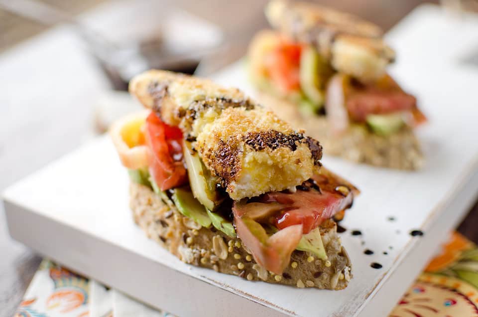 Two slices of seeded bread topped with avocado, tomato, vegetables, and a piece of crispy cheese, drizzled with balsamic glaze on a white plate.