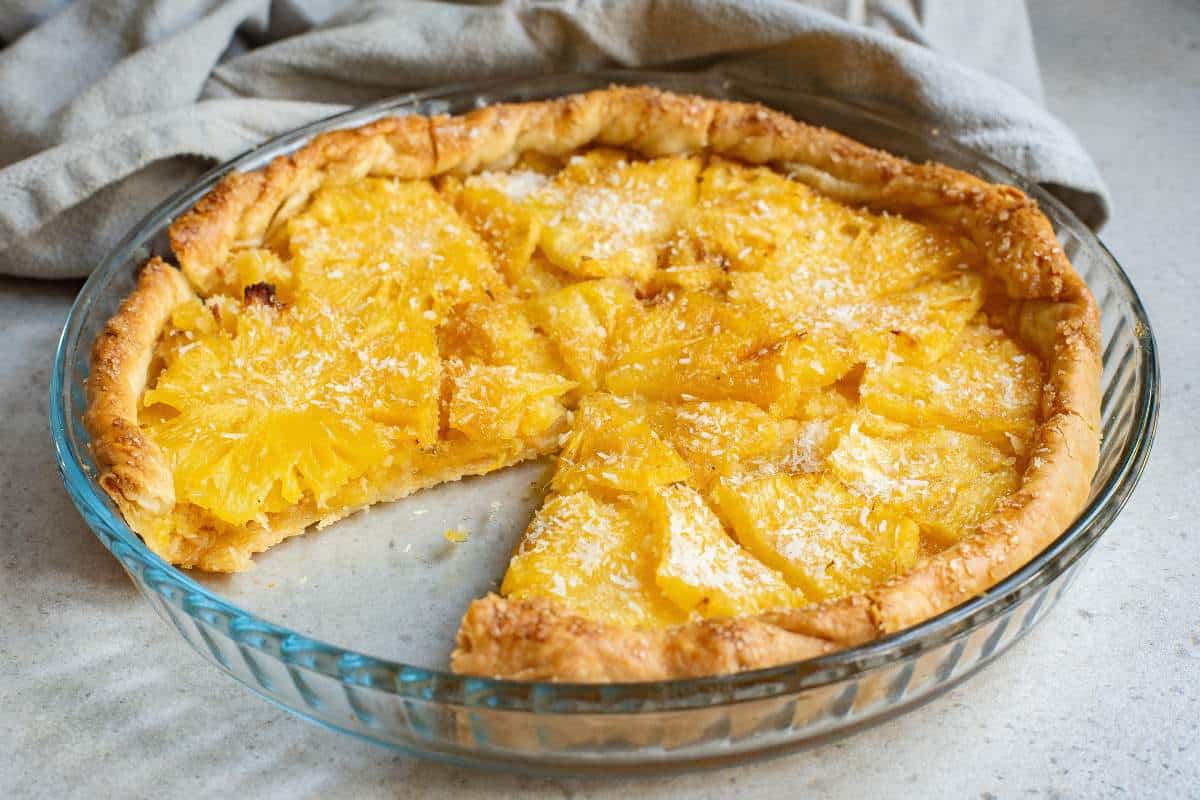 A glass pie dish with a baked pineapple tart, one slice missing, on a light surface with a gray cloth nearby.