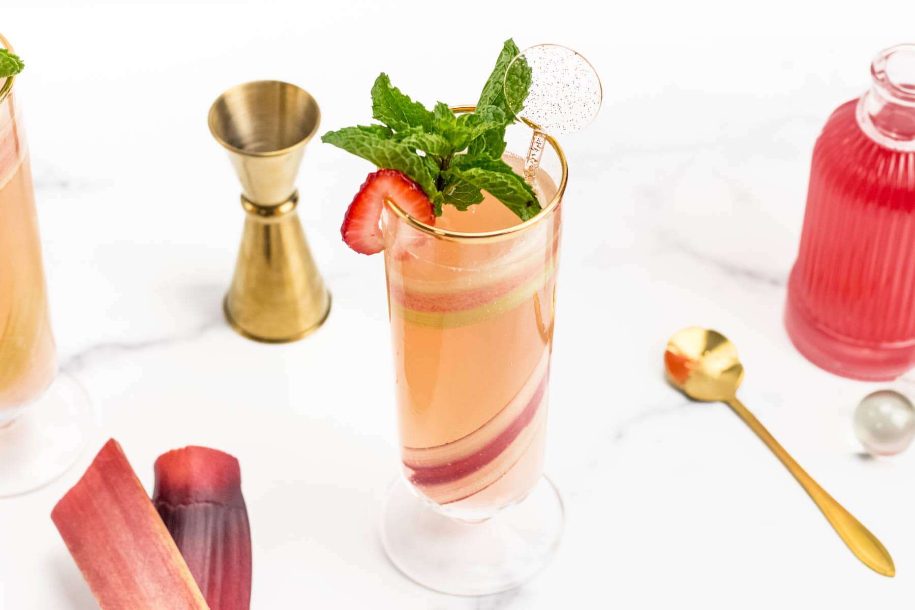 A tall glass of pink cocktail garnished with a mint sprig and strawberry slice, surrounded by a jigger, spoon, and slices of rhubarb on a white surface.
