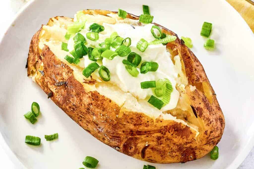 A baked potato topped with sour cream and chopped green onions on a white plate.