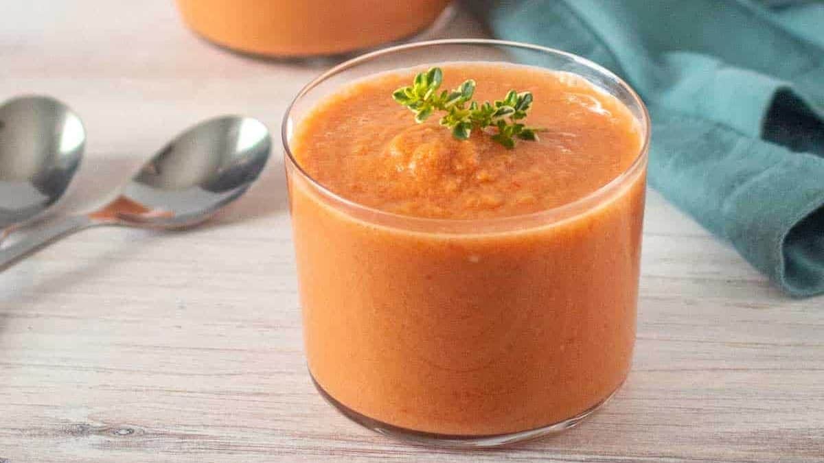 Two glass cups filled with orange-colored soup garnished with greens on a wooden surface. Two spoons and a green cloth are placed near the cups. White tile background.