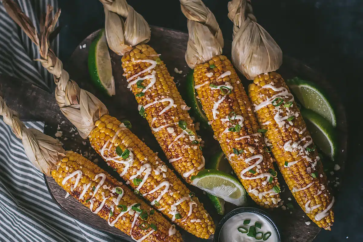 Four ears of grilled corn with braided husks are garnished with a creamy sauce, cheese, and green herbs, served on a dark plate with lime wedges and a small bowl of dip.