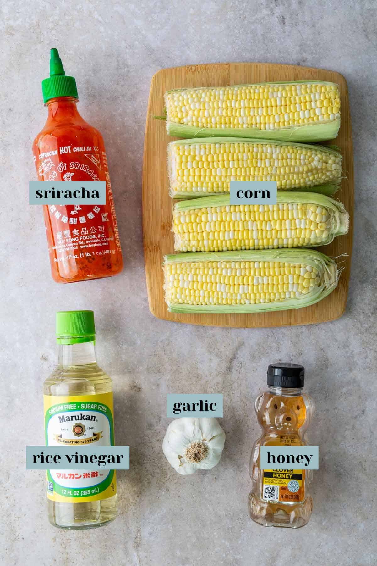 A bottle of sriracha, five ears of corn on a cutting board, a bottle of rice vinegar, a head of garlic, and a bottle of honey arranged on a gray surface.