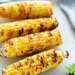 Four grilled corn on the cob with char marks on a cutting board, garnished with fresh parsley.