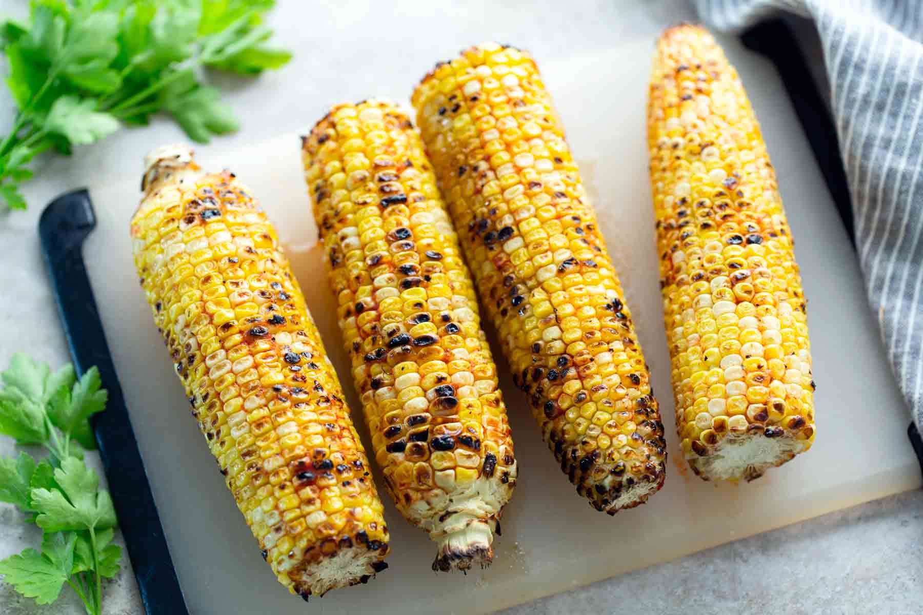 Four grilled ears of corn on a cutting board, with some parsley and a striped cloth napkin nearby.