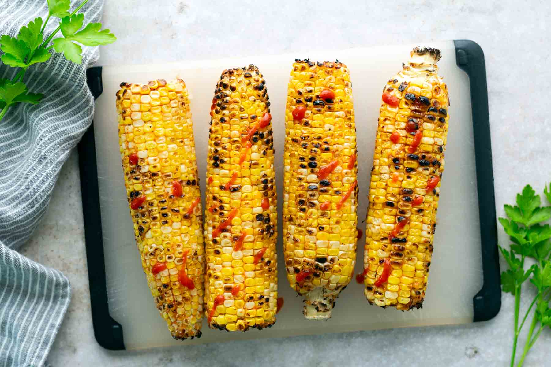 Four grilled corn cobs with seasoning and grill marks placed on a white cutting board, garnished with herbs on the side.
