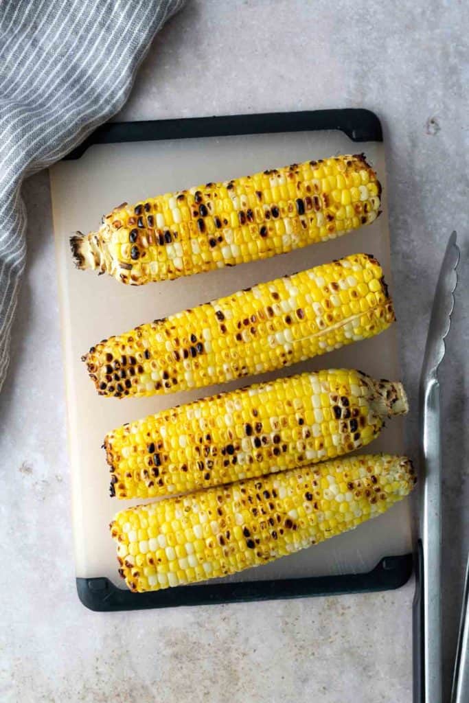 Four grilled corn on the cobs are arranged in a row on a cutting board, with metal tongs placed nearby and a striped cloth partially visible in the corner.