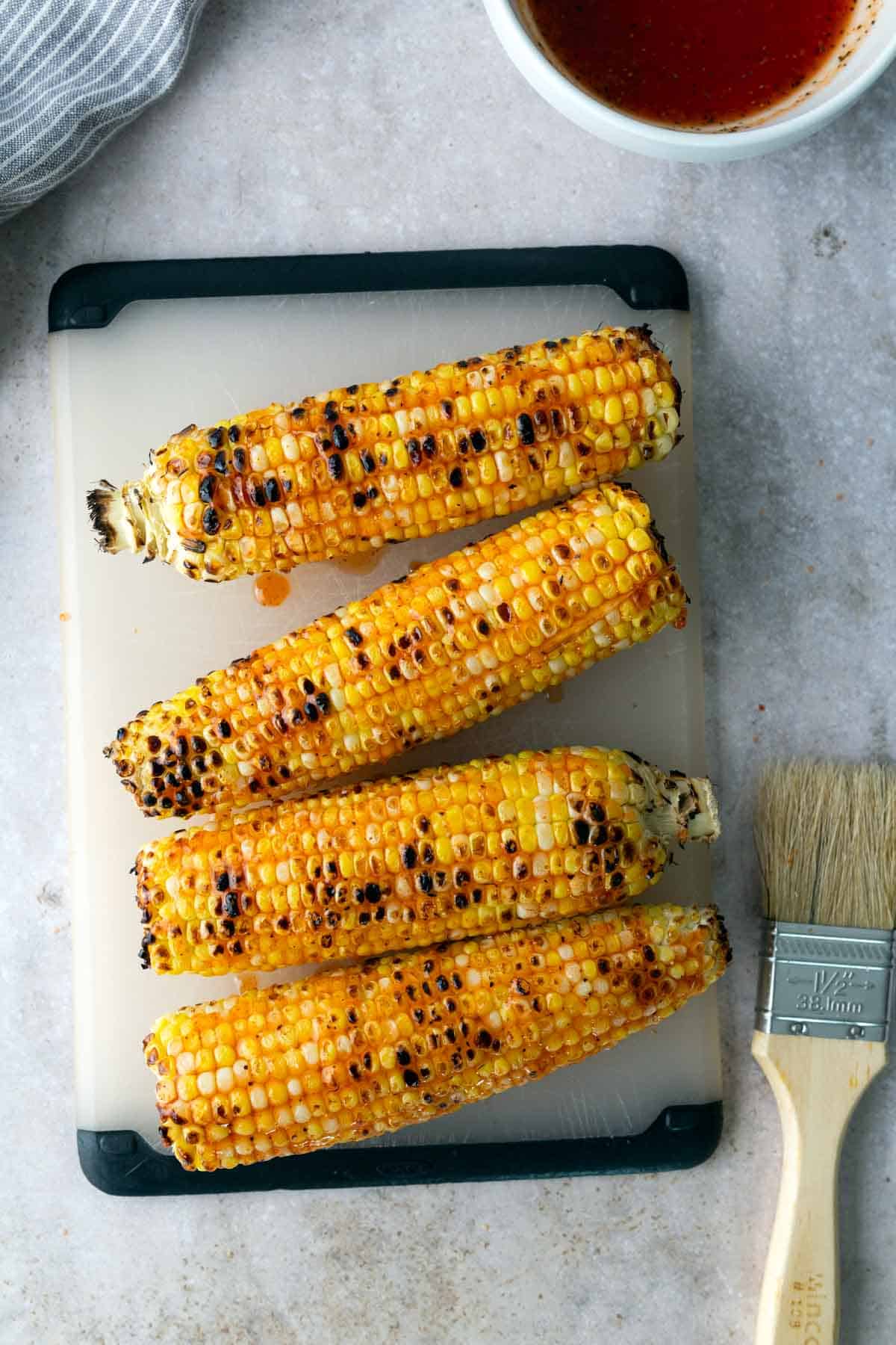 Four grilled corn cobs on a cutting board, with a basting brush and bowl of reddish sauce nearby.