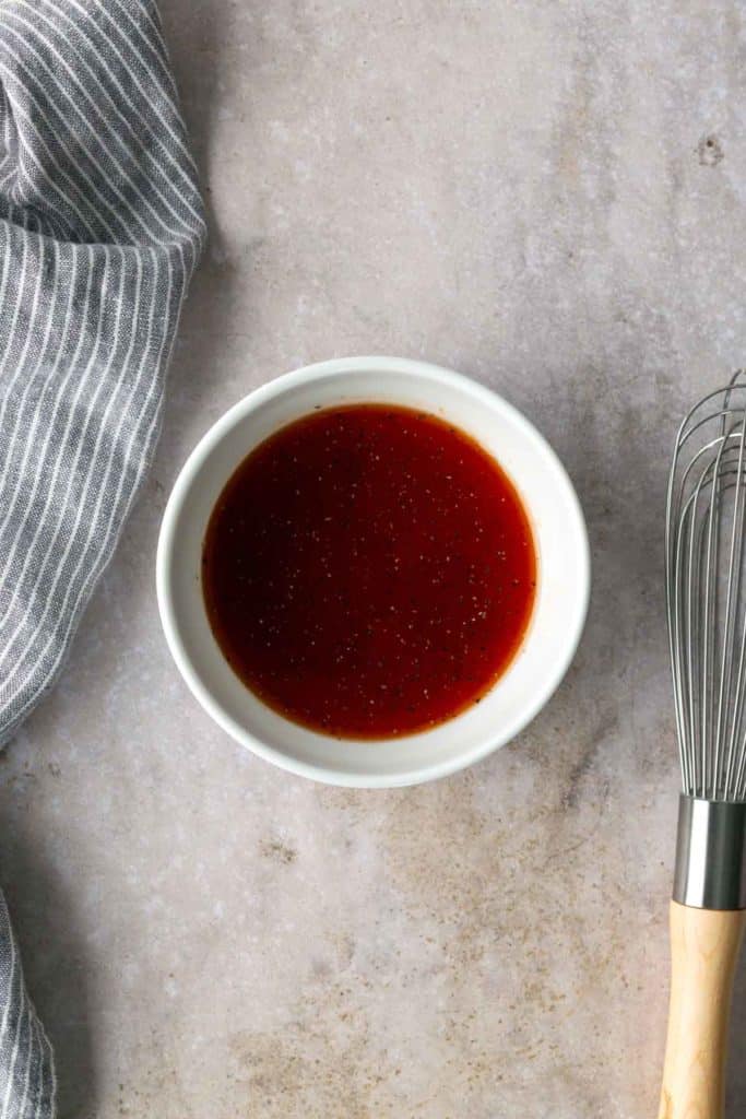 A white bowl filled with reddish-brown sauce sits on a light countertop next to a metal whisk and a gray striped cloth.