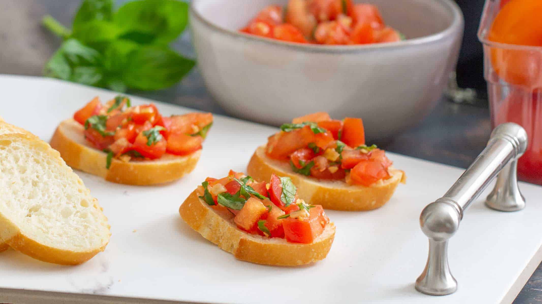 Three slices of baguette topped with diced tomatoes and basil sit on a white cutting board; a bowl of tomato mixture and basil leaves are in the background.