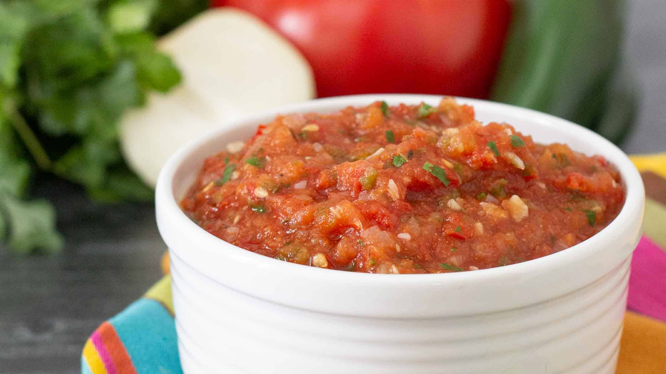 A white bowl filled with chunky red salsa sits on a table; fresh vegetables are blurred in the background.