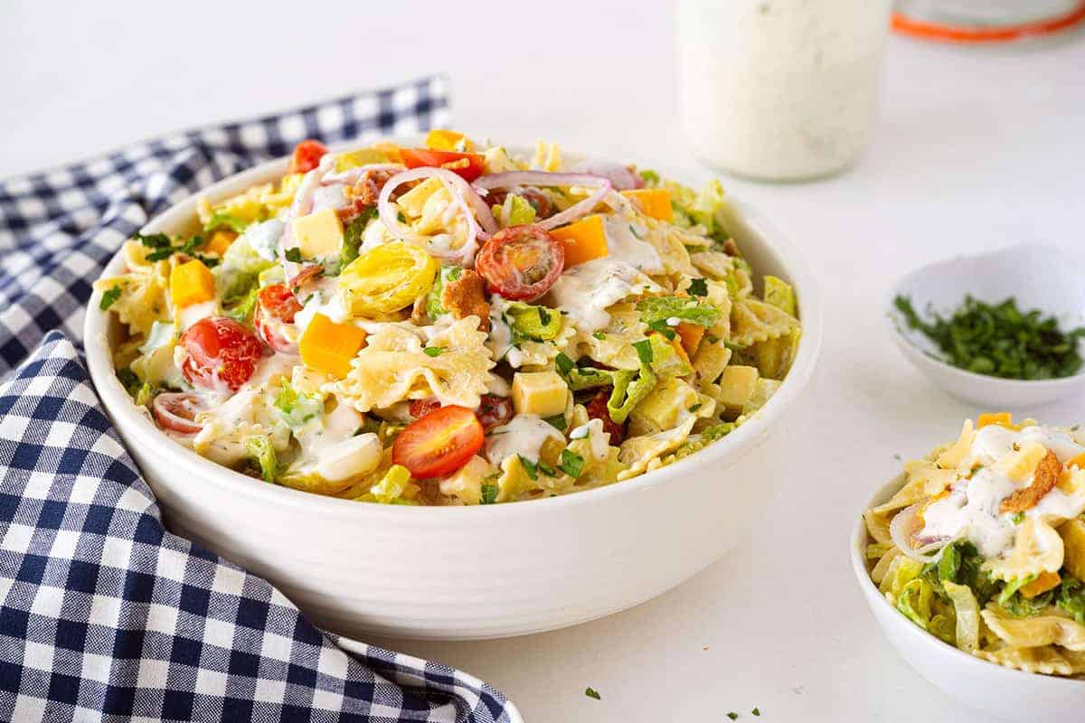 A bowl of pasta salad with cherry tomatoes, lettuce, cheese cubes, red onion, croutons, and creamy dressing, placed on a checkered cloth.