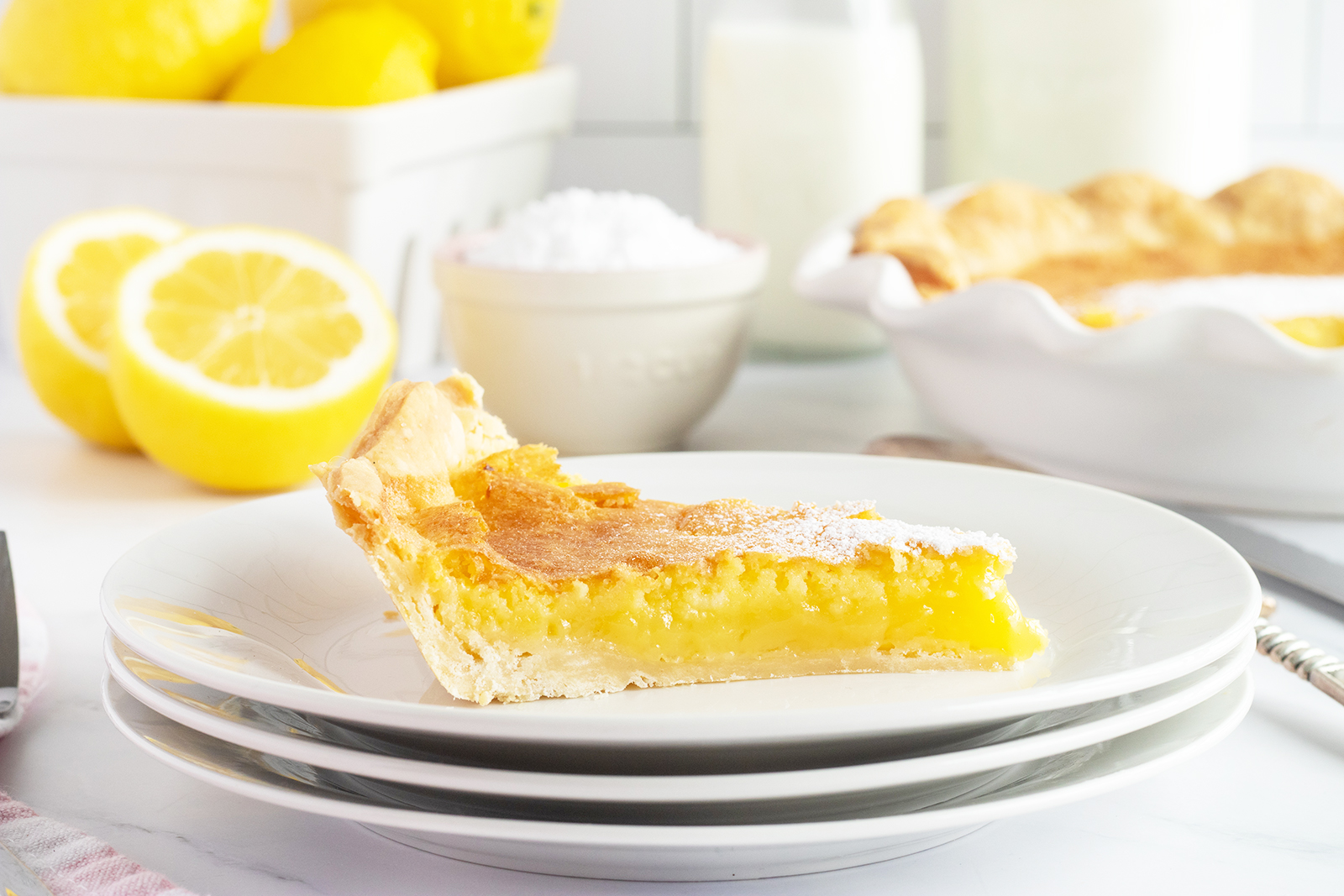 A slice of lemon pie on a stack of white plates, with fresh lemons and a whole pie in the background.