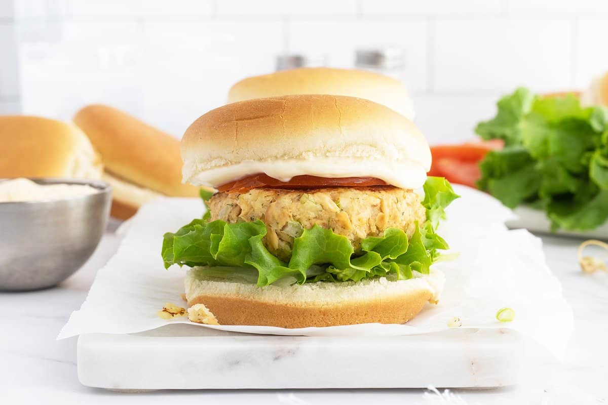 A sandwich with a veggie patty, leaf lettuce, tomato slice, and mayonnaise on a hamburger bun, placed on parchment paper with ingredients in the background.