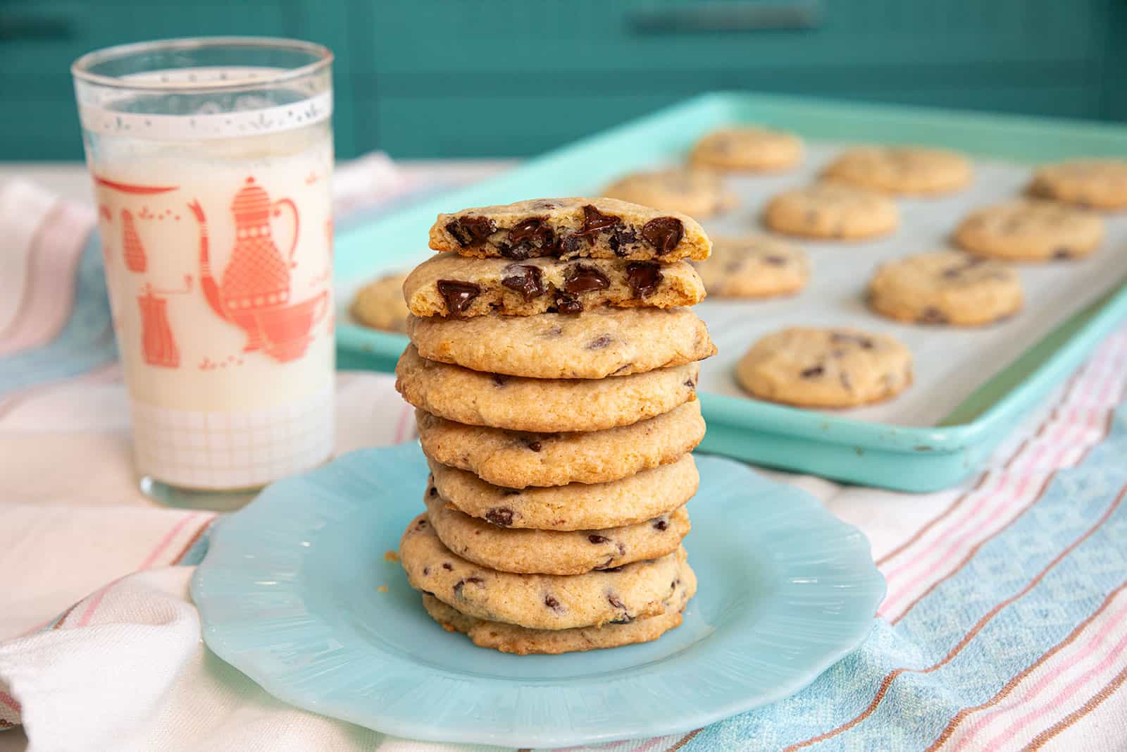 A stack of chocolate chip cookies on a blue plate, with a glass of milk and a tray of more cookies in the background.