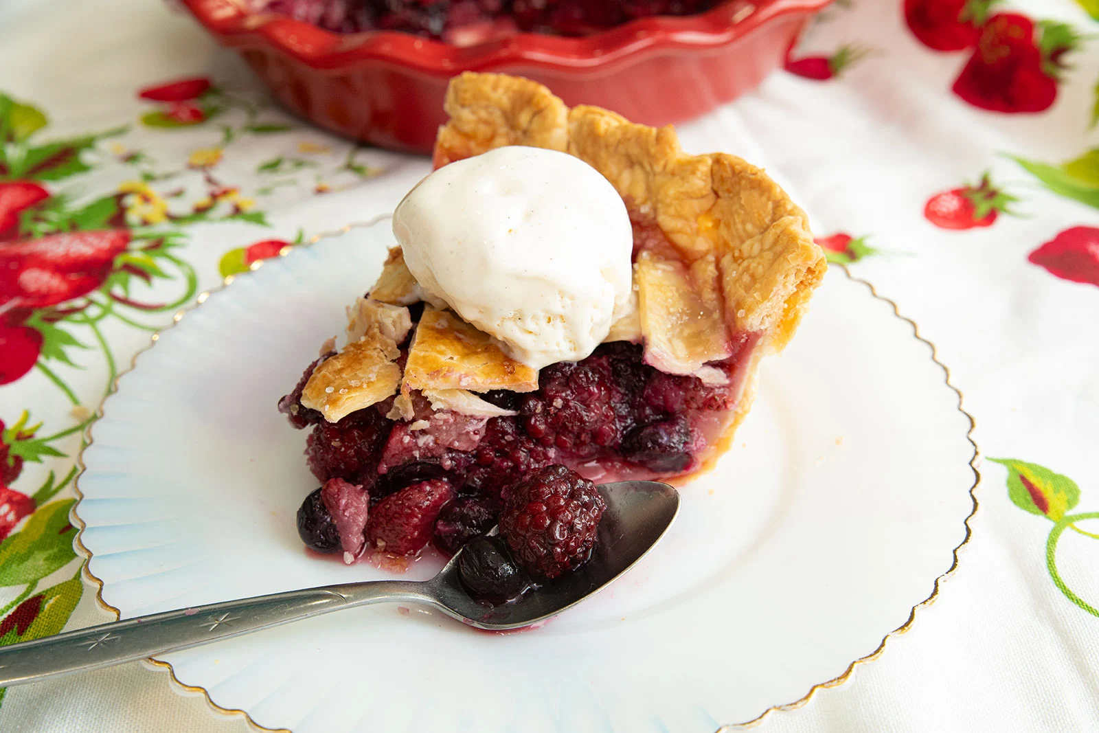 A slice of berry pie topped with a scoop of vanilla ice cream sits on a white plate with a spoon, with the rest of the pie in the background.
