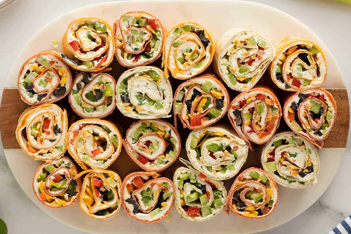 A plate of assorted pinwheel wraps filled with various vegetables, cream cheese, and sliced deli meats, arranged in rows on a white serving platter.