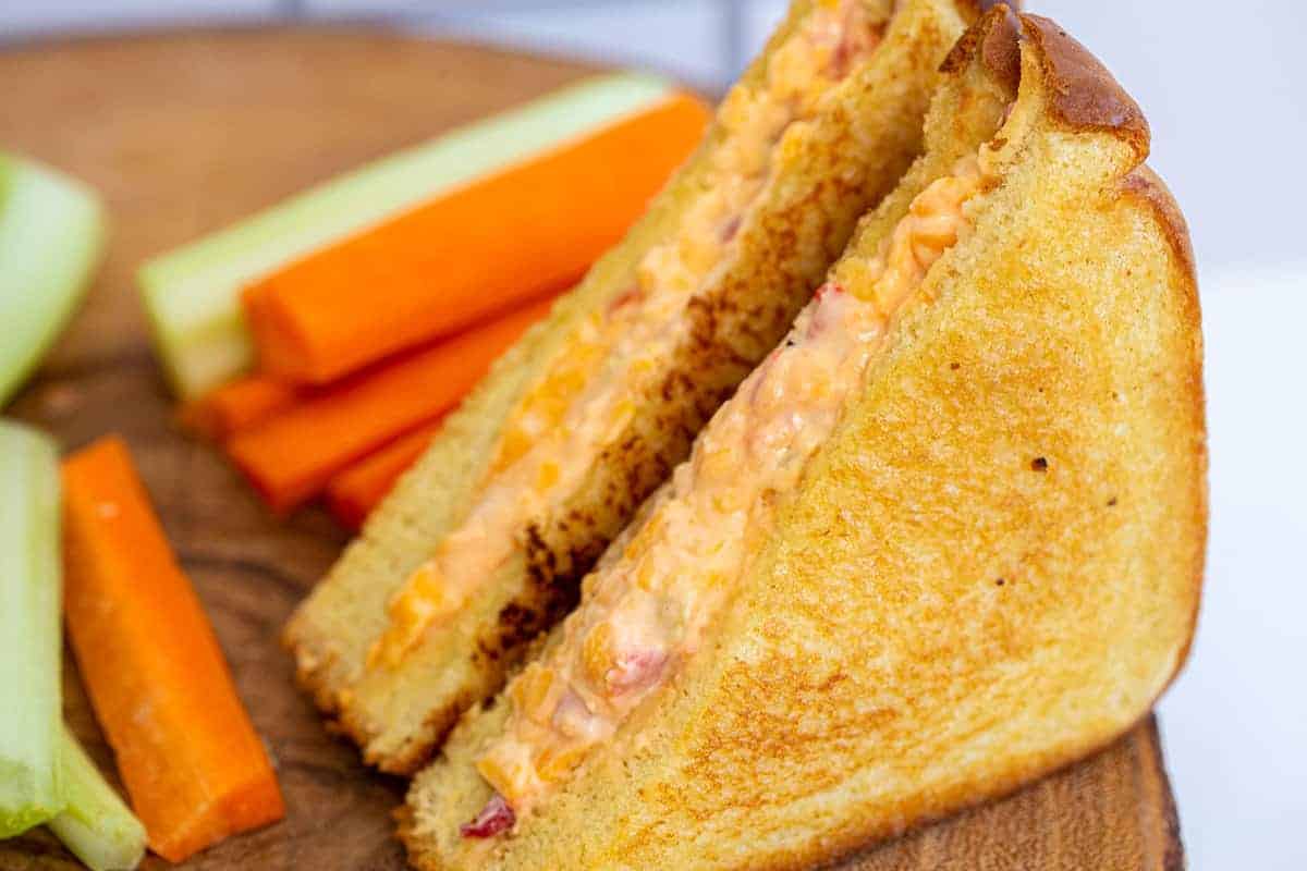 A grilled sandwich with a creamy pimento cheese filling is cut in half and served on a wooden surface with carrot and celery sticks.