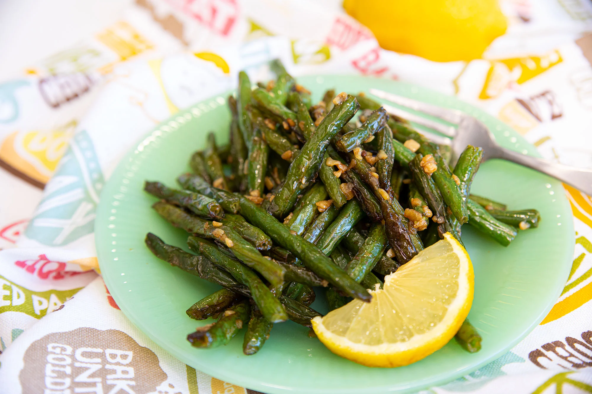 Green beans sautéed with garlic served on a green plate with a lemon wedge, fork beside, and a patterned napkin in the background.