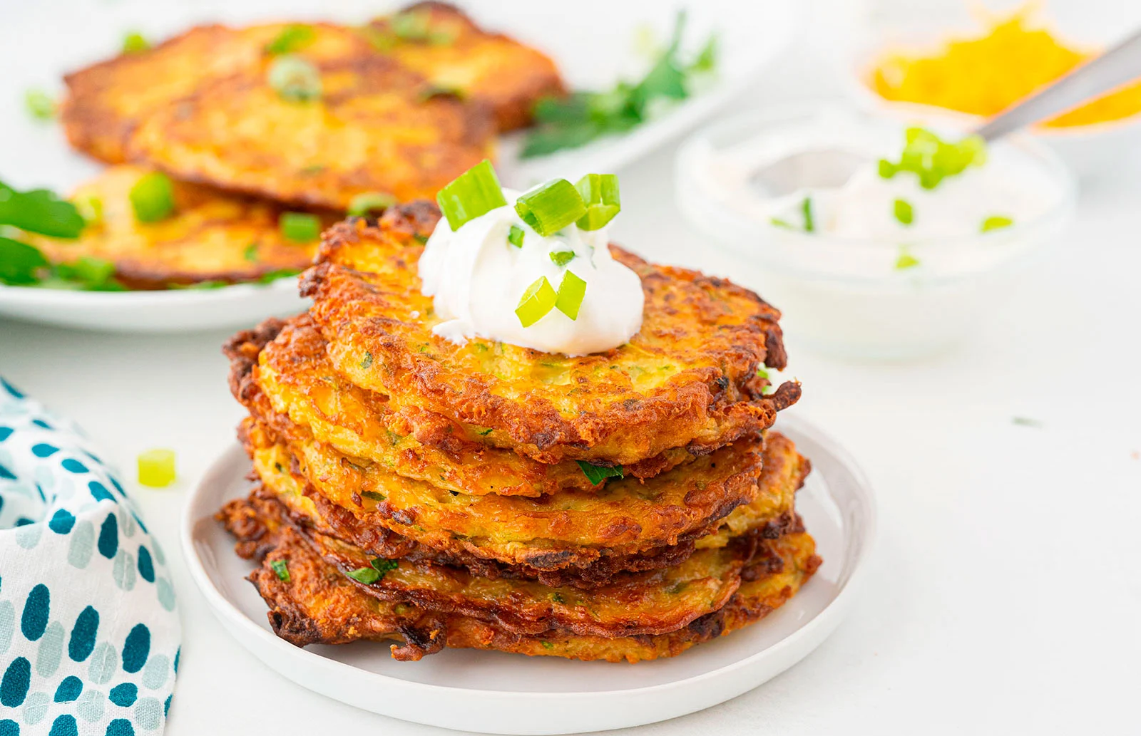A stack of crispy vegetable fritters topped with sour cream and chopped green onions sits on a white plate with herbs and sauce in the background.