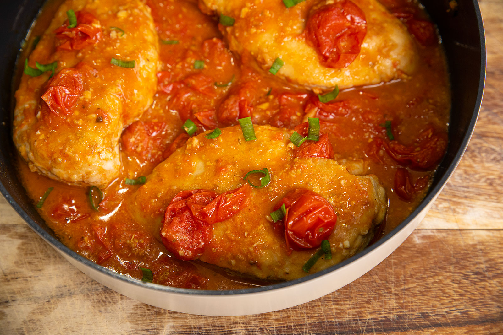 Chicken breasts simmering in a tomato sauce with cherry tomatoes and garnished with chopped green herbs in a saucepan on a wooden surface.